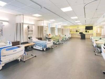 A hospital ward with multiple empty hospital beds, each separated by curtains, medical equipment, and monitors, in a brightly lit, clean environment.