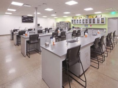 A modern science classroom with multiple lab stations, black chairs, sinks, and lab supplies, under bright ceiling lights. Shelves and a large monitor are visible at the far end.