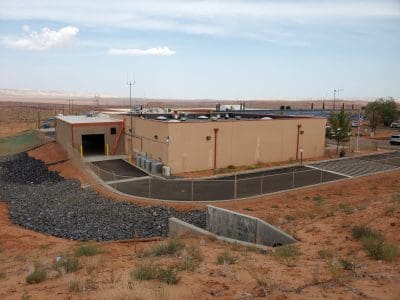 A low, rectangular beige building with rooftop equipment is surrounded by a fence, set in a dry, desert landscape with rocky ground and sparse vegetation.