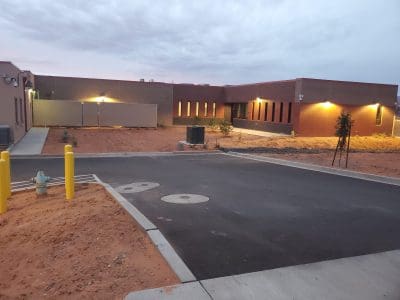 A modern, single-story brick building with illuminated exterior lights, surrounded by a paved road, desert landscaping, and utility features such as a fire hydrant and air conditioning units.
