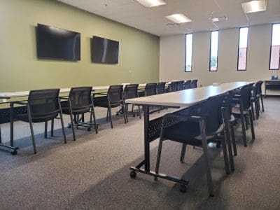 A modern classroom with rows of empty desks and chairs, two wall-mounted monitors, and tall windows letting in natural light.