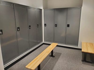 A locker room with gray metal lockers lining two walls and two wooden benches on a carpeted floor.