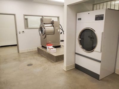 A clean, industrial laundry room with a large stainless steel washing machine, a white dryer, and cleaning supplies on the floor.
