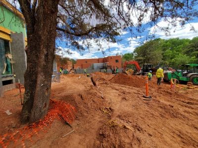 Construction site with workers, machinery, and earthmoving equipment; a tree in the foreground; orange safety fencing along dug-up soil.