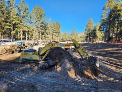 A yellow excavator moves dirt at a construction site surrounded by tall pine trees, with a loader visible in the background under a clear blue sky.