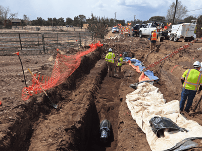 Construction workers in safety gear work in and around a deep trench with a large pipe, surrounded by equipment, vehicles, and orange safety fencing on a dirt site.