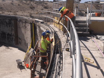 Two workers on a lift and one on stairs perform maintenance or repairs on the curved edge of a large outdoor concrete structure under clear skies.
