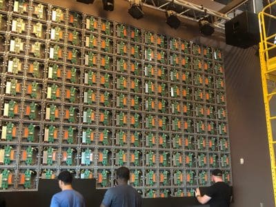 Four people work in front of a large, wall-mounted grid of exposed LED screen panels and circuit boards, with stage lights and scaffolding nearby.