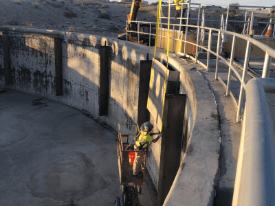 Two construction workers install equipment inside a large, empty circular concrete structure using a crane and a lift.