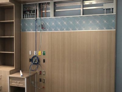 A hospital room wall under construction with exposed wiring, medical gas outlets, and unfinished paneling; a mobile cart is placed nearby.