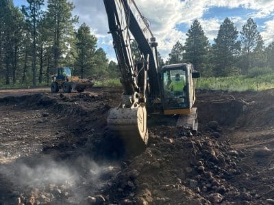 An excavator is digging and moving rocks and soil at a construction site surrounded by trees, with another construction vehicle in the background.