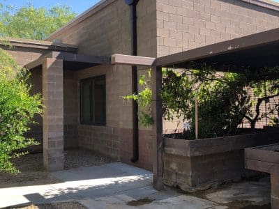 A modern building with tan cinderblock walls, large windows, a covered patio, and a concrete walkway next to plants and gravel under a clear blue sky.
