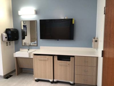 A hospital or clinic room features a sink, countertop, storage cabinets, a wall-mounted TV, soap dispensers, and a mirror against blue and white walls.