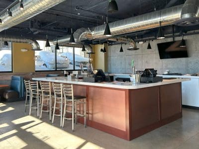 A modern restaurant interior with a central bar, metal chairs, exposed ductwork, concrete walls, large windows, and sunlight streaming in.