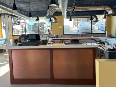 A modern bar counter with a cash register, drink supplies, and overhead lighting in a well-lit space with large windows and visible ductwork.