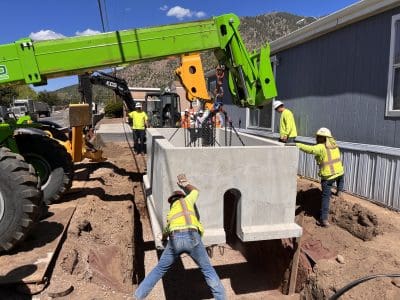 Construction workers guide a large concrete structure being lowered by a green crane into a trench next to a building on a sunny day.