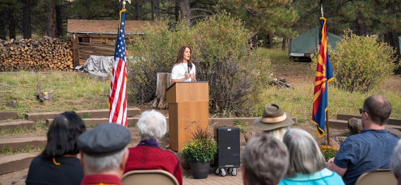 A woman stands at a podium speaking outdoors to a seated audience, with American and Arizona flags on either side and trees in the background, as Kinney celebrates Earth Day—Building Better Together for a Greener Tomorrow.
