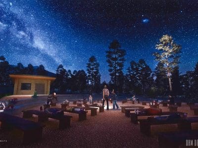 People sit and lie on benches in an outdoor amphitheater under a star-filled night sky surrounded by trees, with a small stage visible on the left.