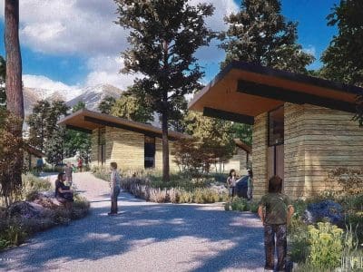 A group of people gather outside modern wooden cabins surrounded by trees and mountains under a partly cloudy sky.