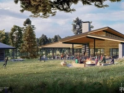 A modern lodge with large windows and sloped roofs sits in a grassy clearing; people gather around an outdoor fire pit and dining area surrounded by trees.