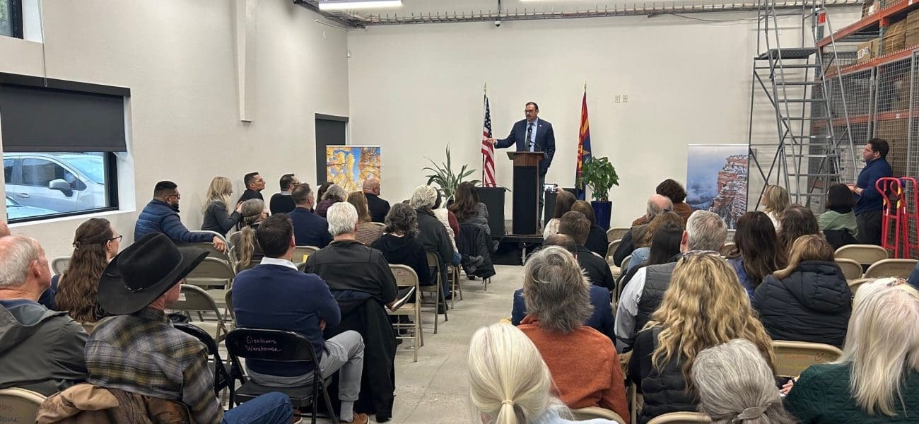 Coconino County Elections Center A man speaks at a podium to an audience in a room with folding chairs, an American flag, and state flag, as banners highlight "Building Better Together for a Greener Tomorrow: How Kinney Honors Earth Day.