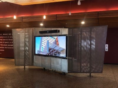 A large screen display in a modern indoor space shows an architectural image; the surroundings feature wood ceiling panels and polished stone flooring.