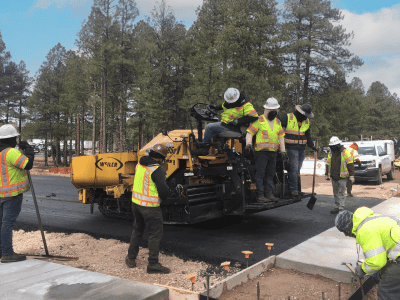 Construction workers in safety gear operate machinery and work on paving a road with fresh asphalt in a wooded area.