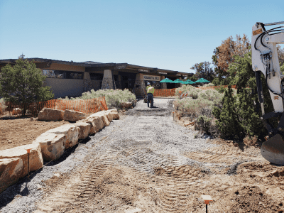 A construction worker levels a gravel path leading to a building entrance marked “Visitor Center,” with large rocks and orange fencing on either side.