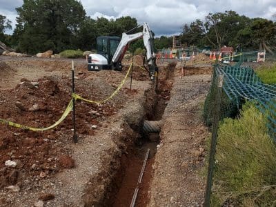 A small excavator digs a trench at a construction site with exposed pipes, safety tape, orange cones, and fencing; cloudy sky overhead.