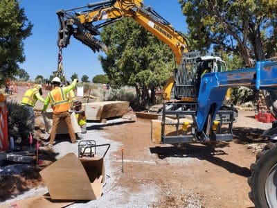 Construction workers in safety vests guide a large stone being lifted by a yellow excavator on a gravel site surrounded by trees.