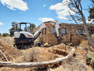 A compact excavator is parked beside a partially damaged stone building, with dirt piles, dry vegetation, and scattered debris in the foreground under a blue sky.