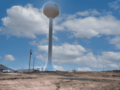 A tall white water tower stands in a fenced area on a dirt lot, with several vehicles and utility poles nearby under a partly cloudy sky.