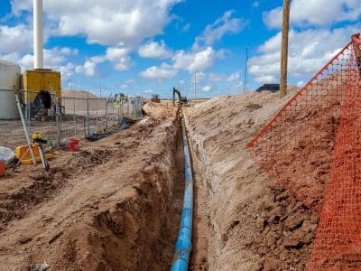 A blue utility pipe is laid in a trench at a construction site, with dirt piles, orange safety fencing, and a water tower in the background under a partly cloudy sky.
