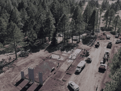 Aerial view of a construction site in a forested area with several vehicles, equipment, and partially built concrete structures.