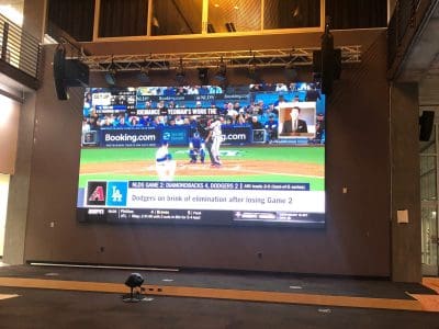 A large indoor screen displays a live baseball game between the Dodgers and Diamondbacks in a spacious, modern room with minimal seating.