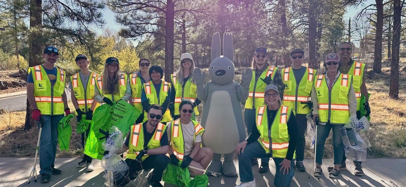 Earth Day A group of people in yellow safety vests pose outdoors with a large gray rabbit mascot, holding green bags, surrounded by trees and sunlight—celebrating Building Better Together for a Greener Tomorrow: How Kinney Honors Earth Day.