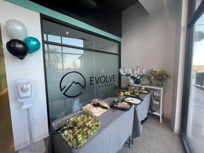 A table with salads and dishes set up for an event in front of a glass wall labeled "Evolve Flagstaff," with balloons and a flower arrangement nearby.