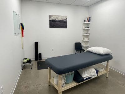 A minimalist medical or therapy room with a massage table, chair, shelving with towels, small weights, a foam roller, and a wall photo.