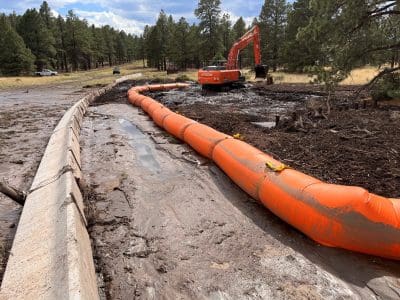 An orange water-filled barrier lines a muddy area near a forest road, with an excavator and pine trees visible in the background.