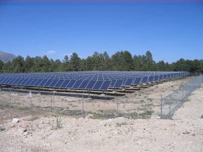 Rows of solar panels, part of the Northern Arizona University PV System, are installed on a fenced-in plot of land with trees and a clear blue sky in the background.