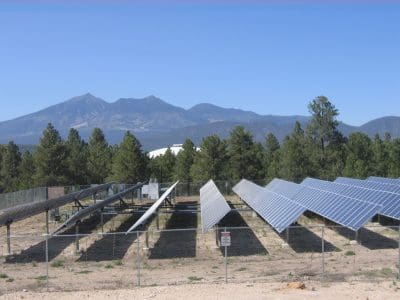 Rows of solar panels from the Northern Arizona University PV System are installed in a fenced area with trees and mountains in the background under a clear blue sky.