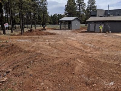 A construction site with dirt ground, a small gray building, two workers, and trees in the background. A sign is posted on the left side of the image.