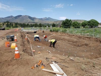 Several construction workers install rebar in a large, excavated dirt pit at an outdoor construction site with mountains in the background.