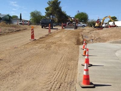 A dirt road under construction with traffic cones, construction vehicles, and equipment visible. The sky is clear and a few trees are in the background.