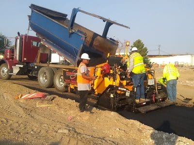Construction workers operate paving machinery as asphalt is poured from a dump truck onto a road under construction at a worksite.
