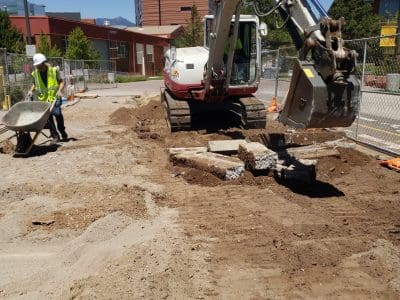A construction worker uses a wheelbarrow while an excavator moves dirt at a construction site with buildings in the background.