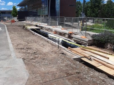 Construction site with exposed trench and wooden planks near a modern building, fenced off for safety on a sunny day.