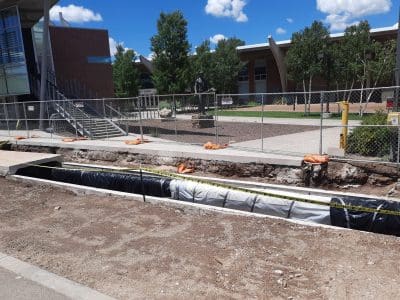 A long trench is dug along a sidewalk on a campus, bordered by construction fencing and caution tape, with buildings and trees in the background.