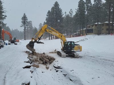 A yellow excavator breaks up and moves rocks on a snow-covered construction site with trees and buildings in the background.
