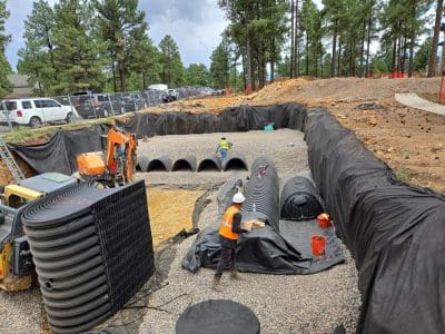 Construction workers install large black plastic drainage pipes and structures in a deep excavation site, surrounded by gravel and lined with black fabric, near a forested area.
